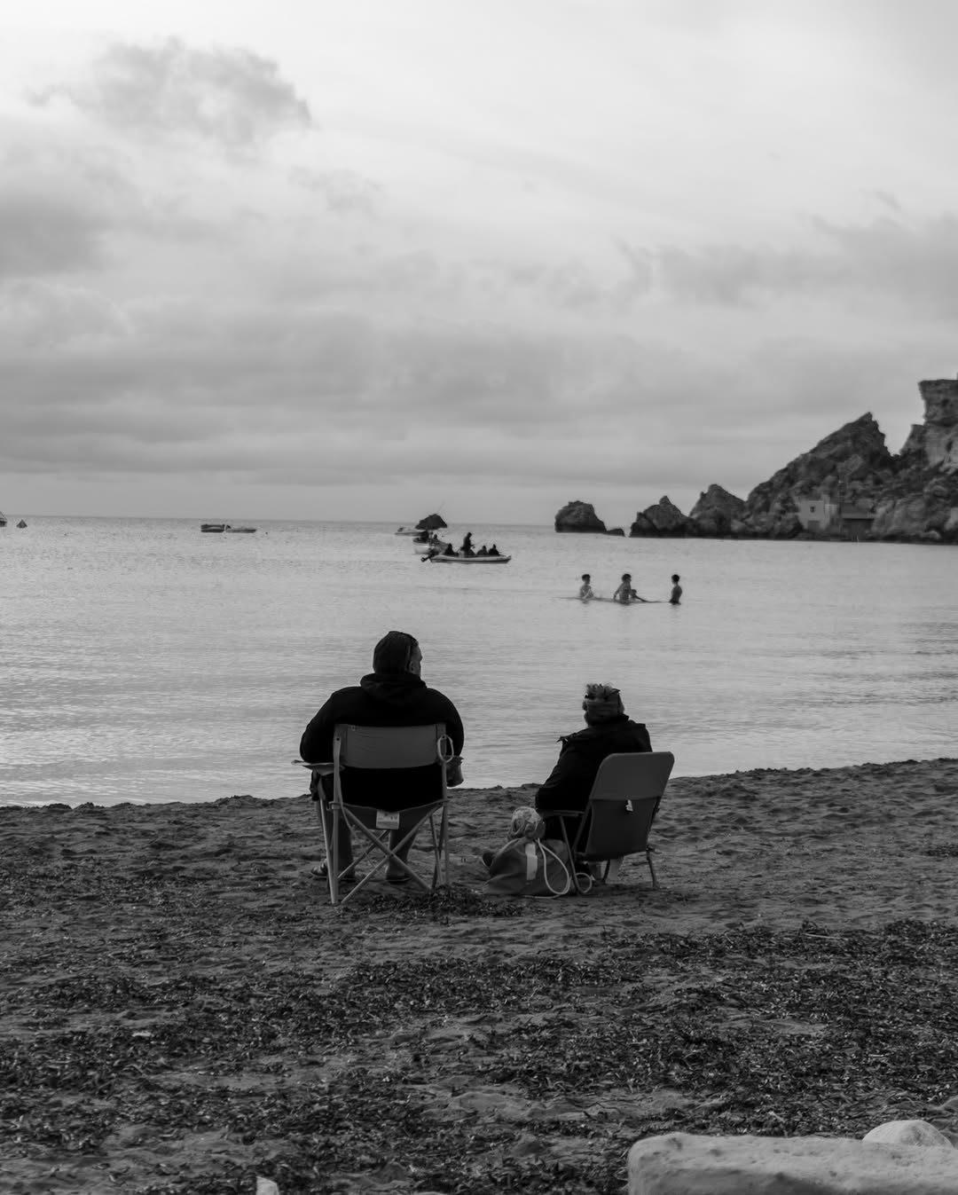 Black and white photo by Chiemeka Madufor: two people in folding chairs on a beach watch swimmers and boats in a calm bay below rocky cliffs